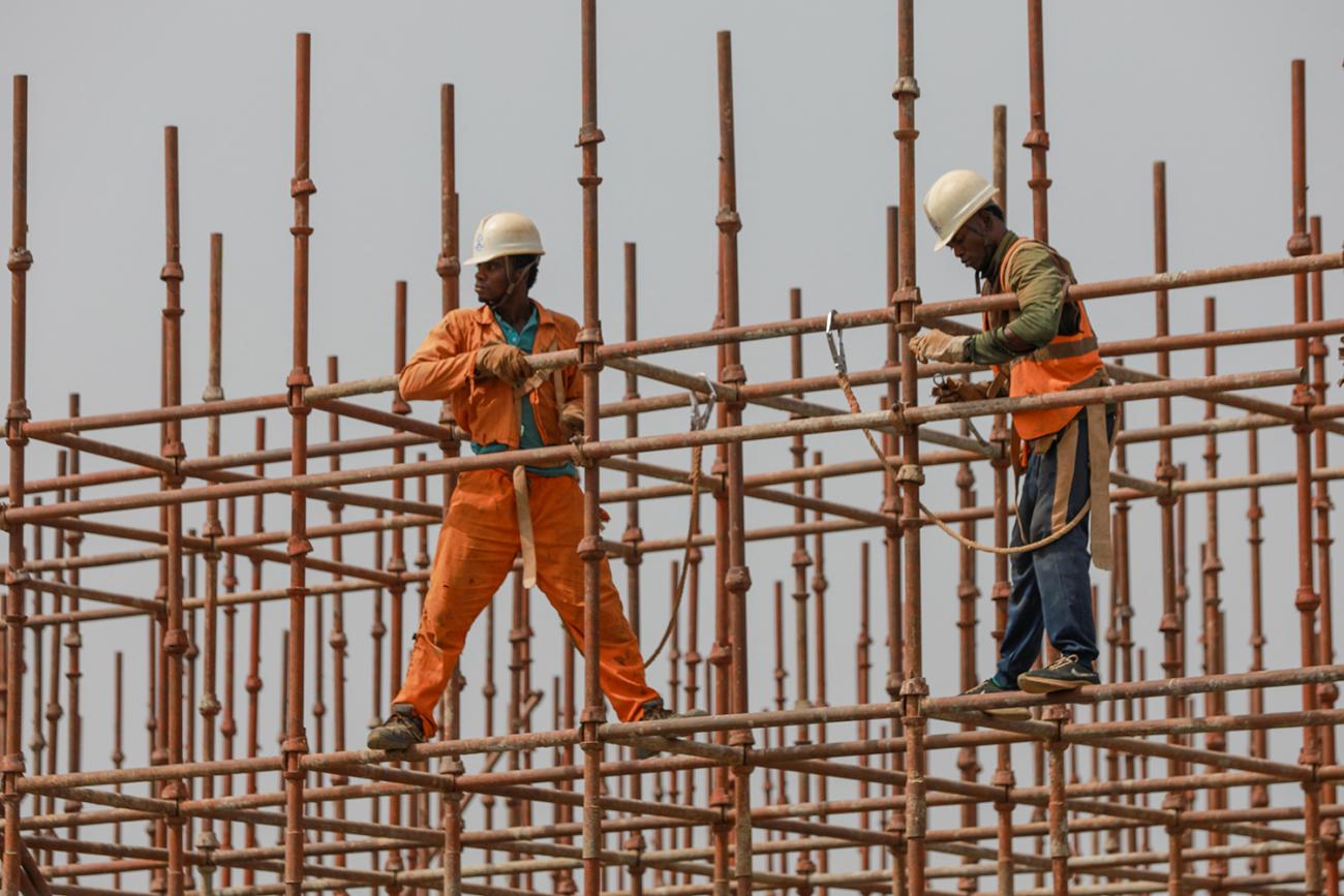 Construction workers stand on a scaffolding on February 18, 2020 in Abuja, Nigeria—one of the countries with a looming crisis of an unmet need for housing coupled with a projected population increase. Picture shows a man in a bright orange jump suit and a yellow contruction hat balancing with his legs wide apart on a metal scaffolding against a grey sky. REUTERS/Afolabi Sotunde