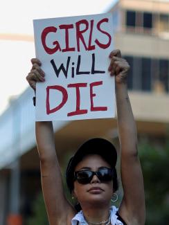 A woman wearing a black sports bra holds above her head  a sign  that reads "girls will die"