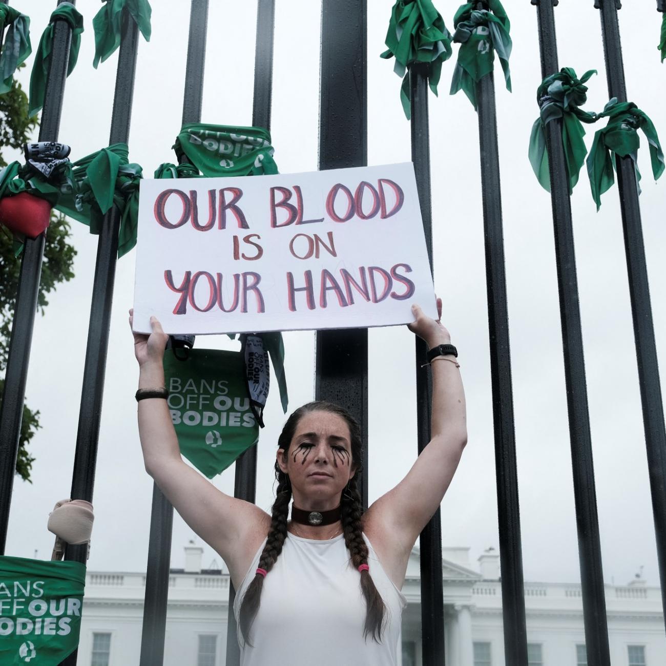 A woman with two braids wearing a white tank top holds up a sign that reads "our blood is on your hands" in front of the white house