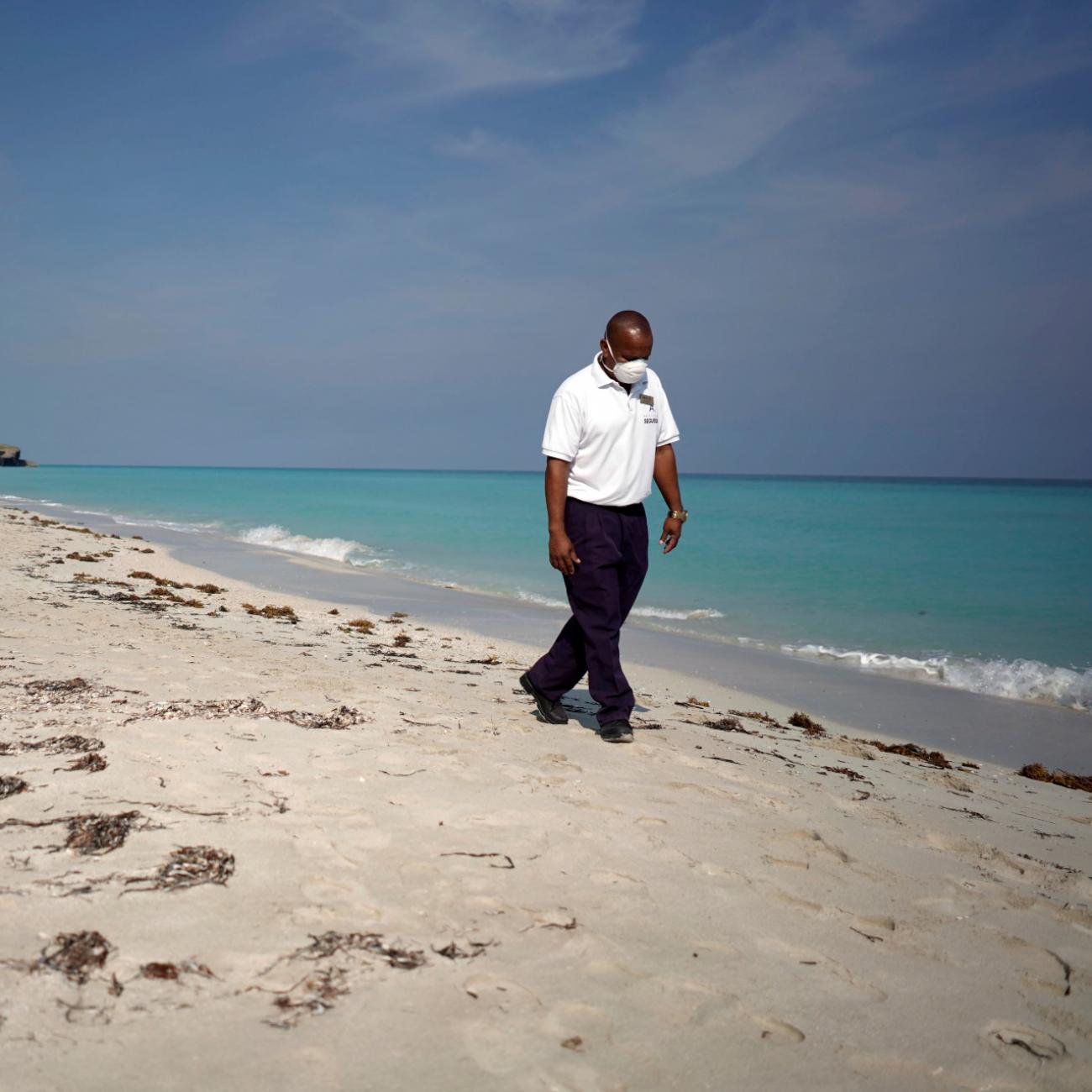A security agent wearing a white protective mask, white polo shirt, and black slacks, walks along the shoreline of a white sand beach with crystal blue waters in Cuba