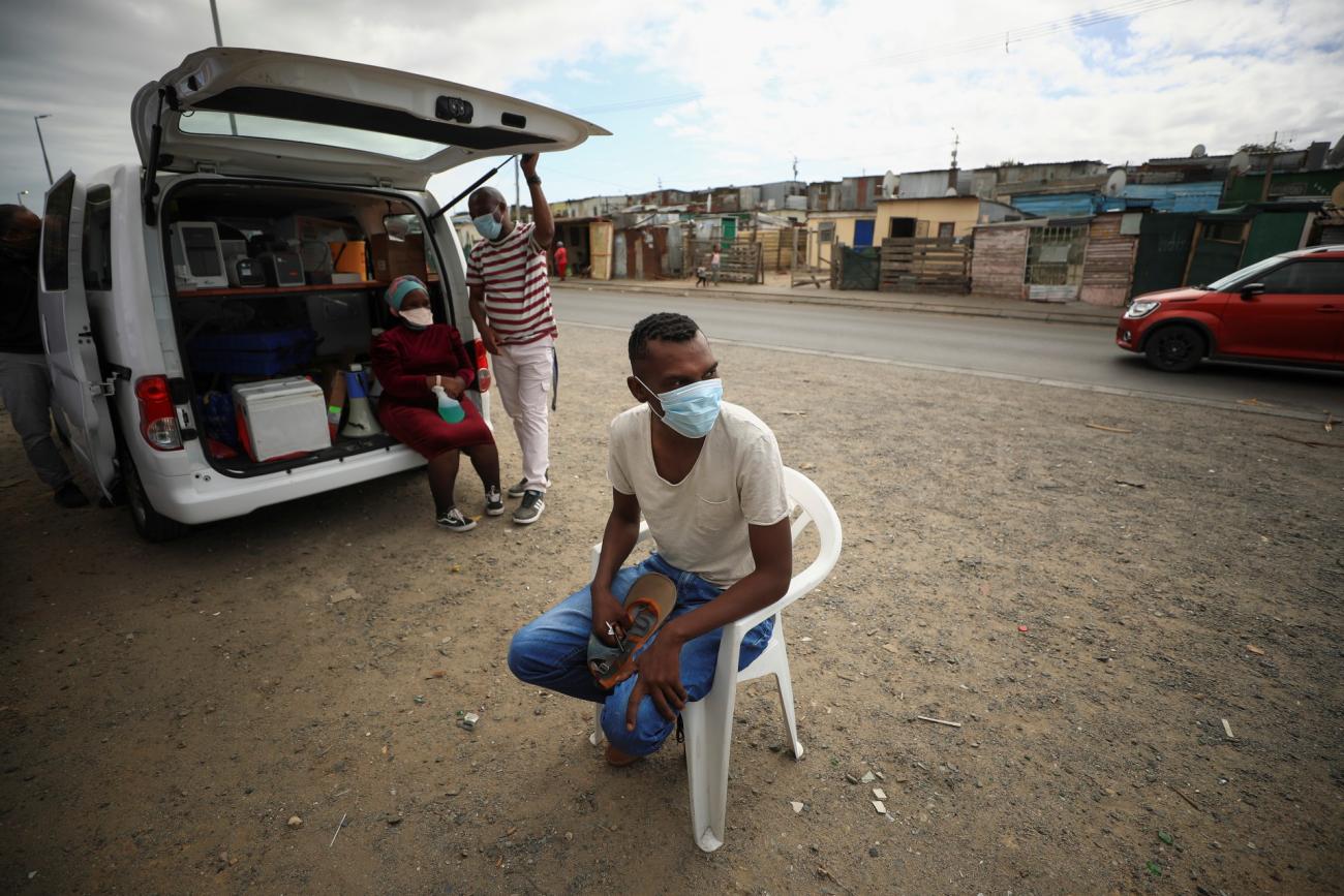 a young man wearing a white t-shirt, blue jeans, and a blue surgical face mask, sits on a plastic chair outside a mobile TB testing clinic