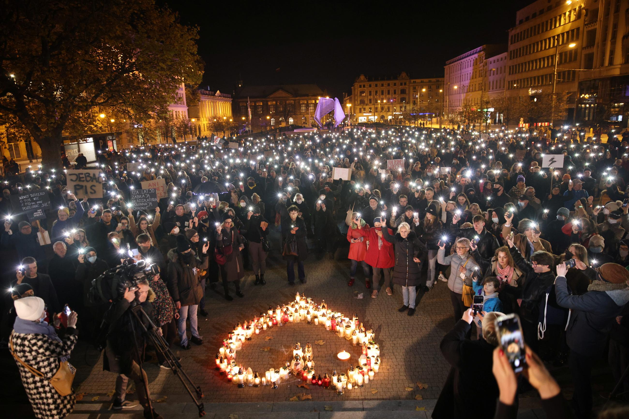 A large crowd of people hold lighted cell-phones at night in a vigil for Izabela, a woman who died after being refused abortion care. On the ground in front of them is a formation of red and white candles in the shape of a heart.